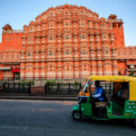 Indian man drives auto rickshaw (tuk-tuk) on streets of Rajasthan, India. Hawa Mahal (The Wind Palace) on the background.